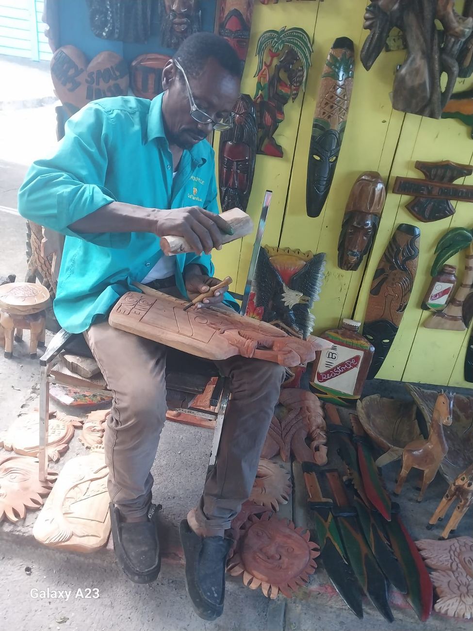 A carver at his stall carving a wooden plaque by hand with a knife, wearing glasses and a Pineapple Craft Market polo shirt, surrounded by carved masks and plaques