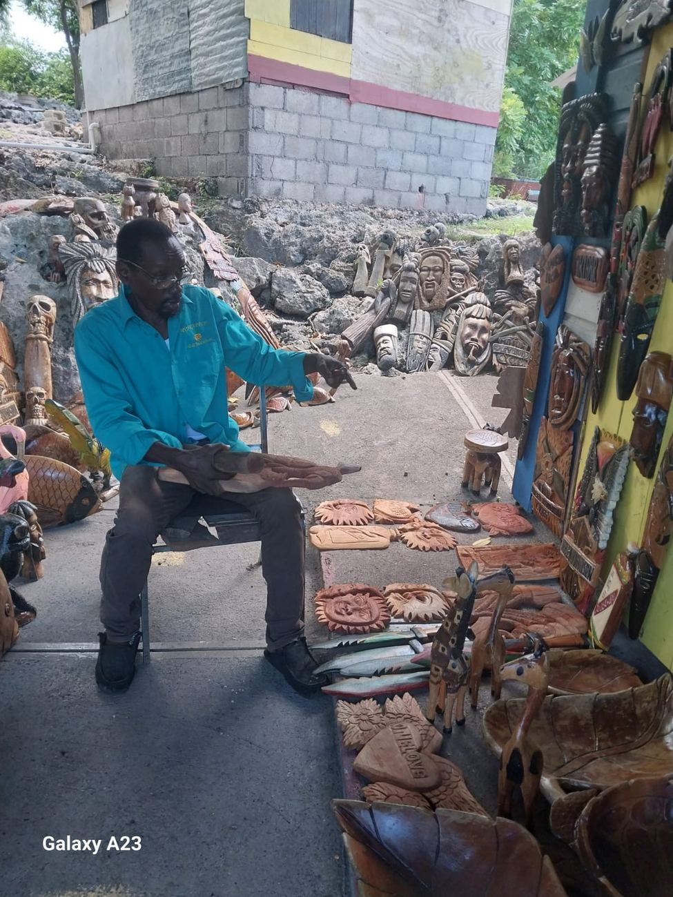 A carver seated outdoors beside a rock wall lined with dozens of finished wood mask carvings laid out to season in the sun