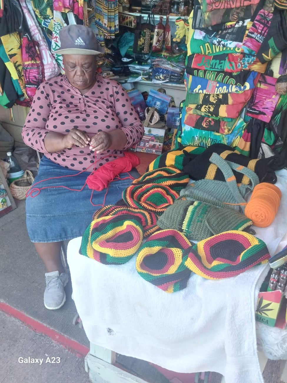 A vendor crocheting a red, gold and green tam at her table, with finished crochet bags, tams and cushions spread out in front of her
